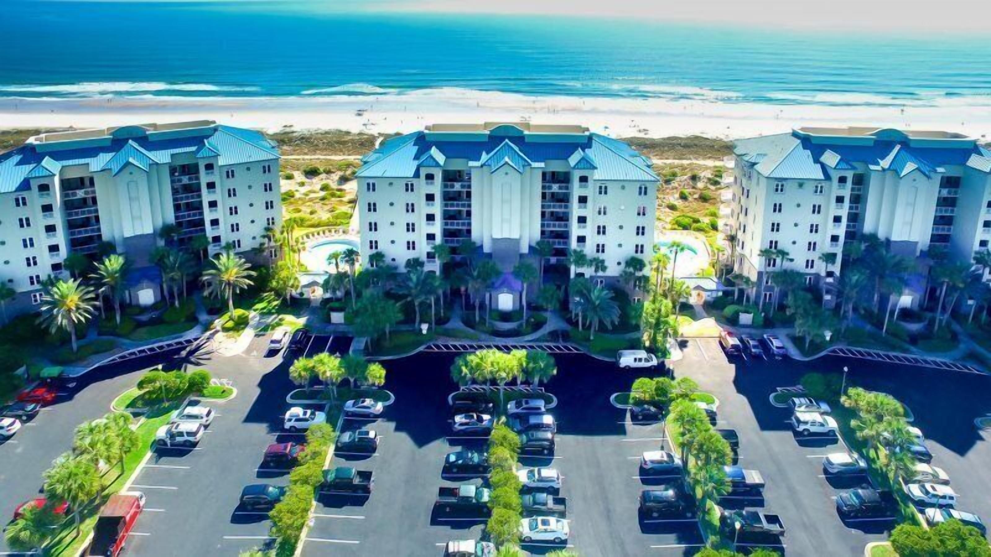 A hotel with multiple buildings, a parking lot, and a beach view.