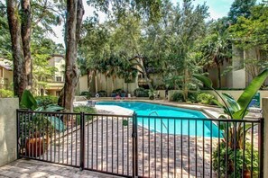 A swimming pool surrounded by a brick patio, with a metal fence and lush greenery.