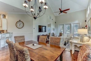 A spacious living room with a wooden dining table, wicker chairs, a chandelier, and a ceiling fan.