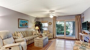 Un salon comprenant un canapé, un fauteuil et une table basse. On y trouve également un ventilateur de plafond, un tableau accroché au mur et une fenêtre avec des rideaux.