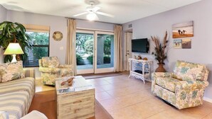 Un salon comprenant un canapé, des fauteuils, une table basse, un téléviseur et une grande fenêtre avec des rideaux.