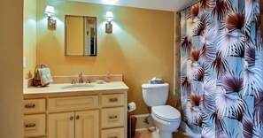 Une salle de bain avec un meuble-lavabo beige, des toilettes blanches et un rideau de douche orné d’un motif de feuilles tropicales.