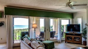 A living room with a sofa, a television, and a balcony with a view of the ocean.