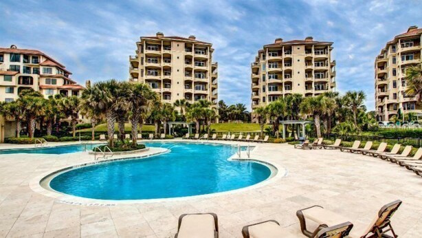 A hotel with a central swimming pool surrounded by lounge chairs and palm trees.