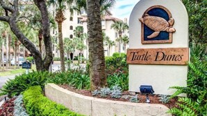 A sign for Turtle Dunes with a turtle graphic, surrounded by palm trees and a landscaped area.