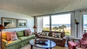 A living room with a green sofa, a wicker sofa, and a round coffee table. There are two framed pictures on the wall, a floor lamp, and a view of the beach through large windows. - 1196-beach-walker (Fernandina Beach)