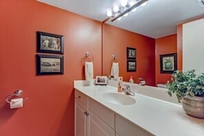 A bathroom with a white sink, two framed pictures on the wall, a mirror, and a potted plant. - 3062-club-villas (Fernandina Beach)