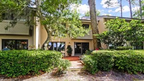 A modern building with a covered patio area, surrounded by well-maintained hedges and trees. - 3062-club-villas (Fernandina Beach)