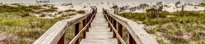 A wooden boardwalk leading to a beach with dunes and sparse vegetation.