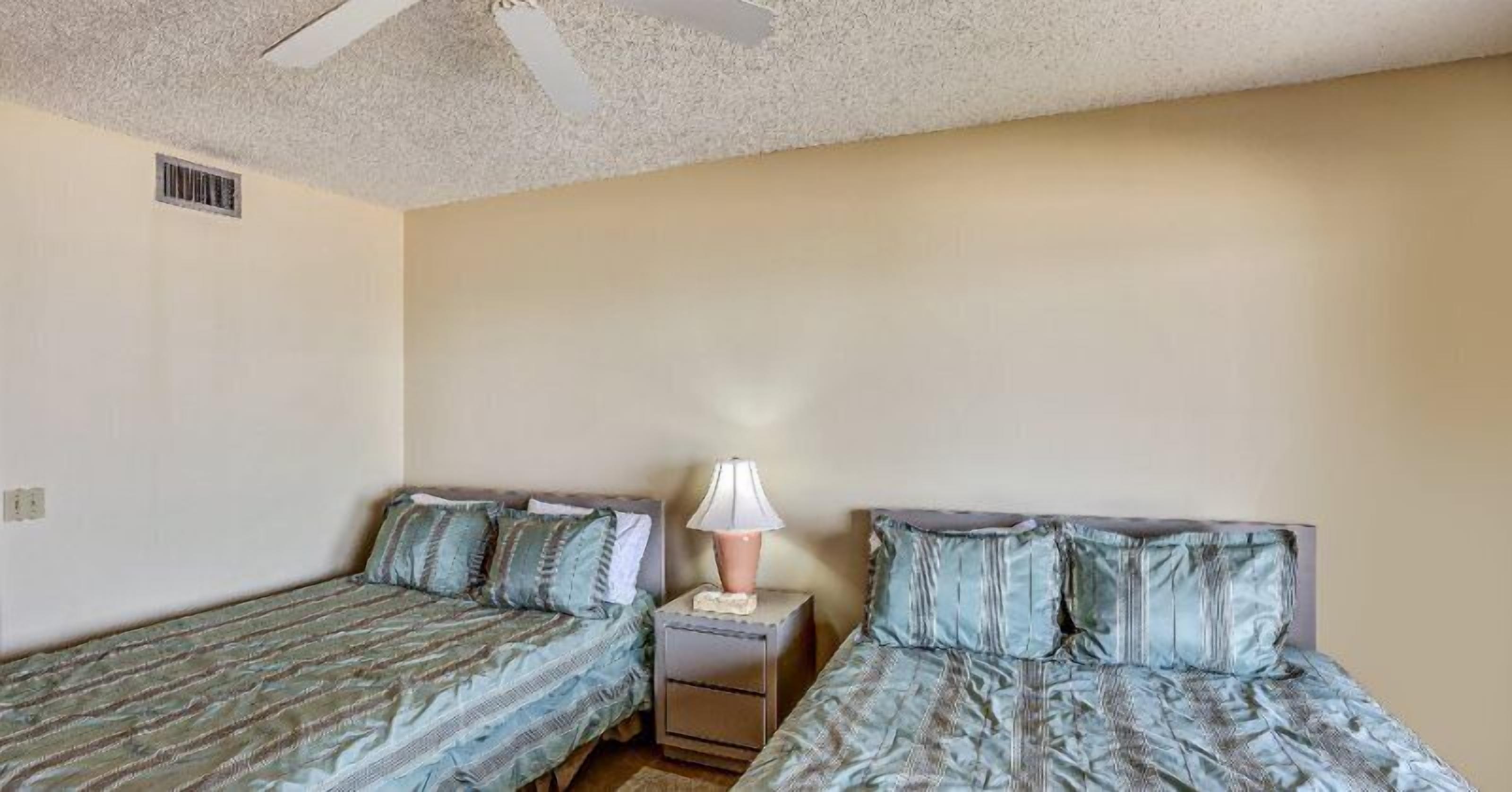 A bedroom with two single beds, a nightstand with a lamp, and a ceiling fan.