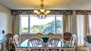 Zona pranzo con tavolo e sedie in vetro, un lampadario e vista sugli alberi attraverso ampie finestre.