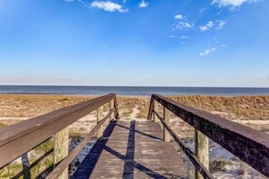 Ein hölzerner Steg führt zu einem Strand mit Dünen und einem klaren Himmel.