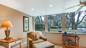 Una habitación acogedora con una butaca cómoda, una mesita de madera con una lámpara y vistas a un jardín a través de ventanas amplias.