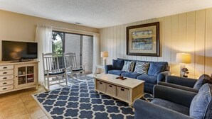 A living room with a TV, a sofa, a coffee table, a dining area with chairs, and a balcony with a view. - 2100-beachwood (Fernandina Beach)