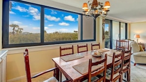 A dining area with a large window, wooden table, chairs, and a chandelier. - 1129 Beach Walker (Fernandina Beach)