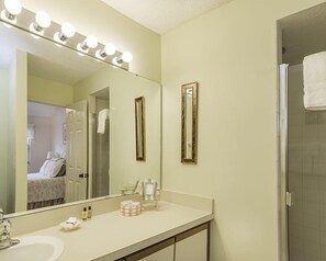 A bathroom with a vanity, a large mirror with lights, two wall mirrors, a shower, and a towel hanging on the door. - 306-sailmaker (Fernandina Beach)