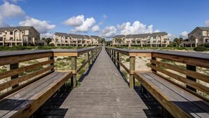 A wooden boardwalk leading to a row of apartment buildings with balconies, surrounded by sand and greenery. - 306-sailmaker (Fernandina Beach)
