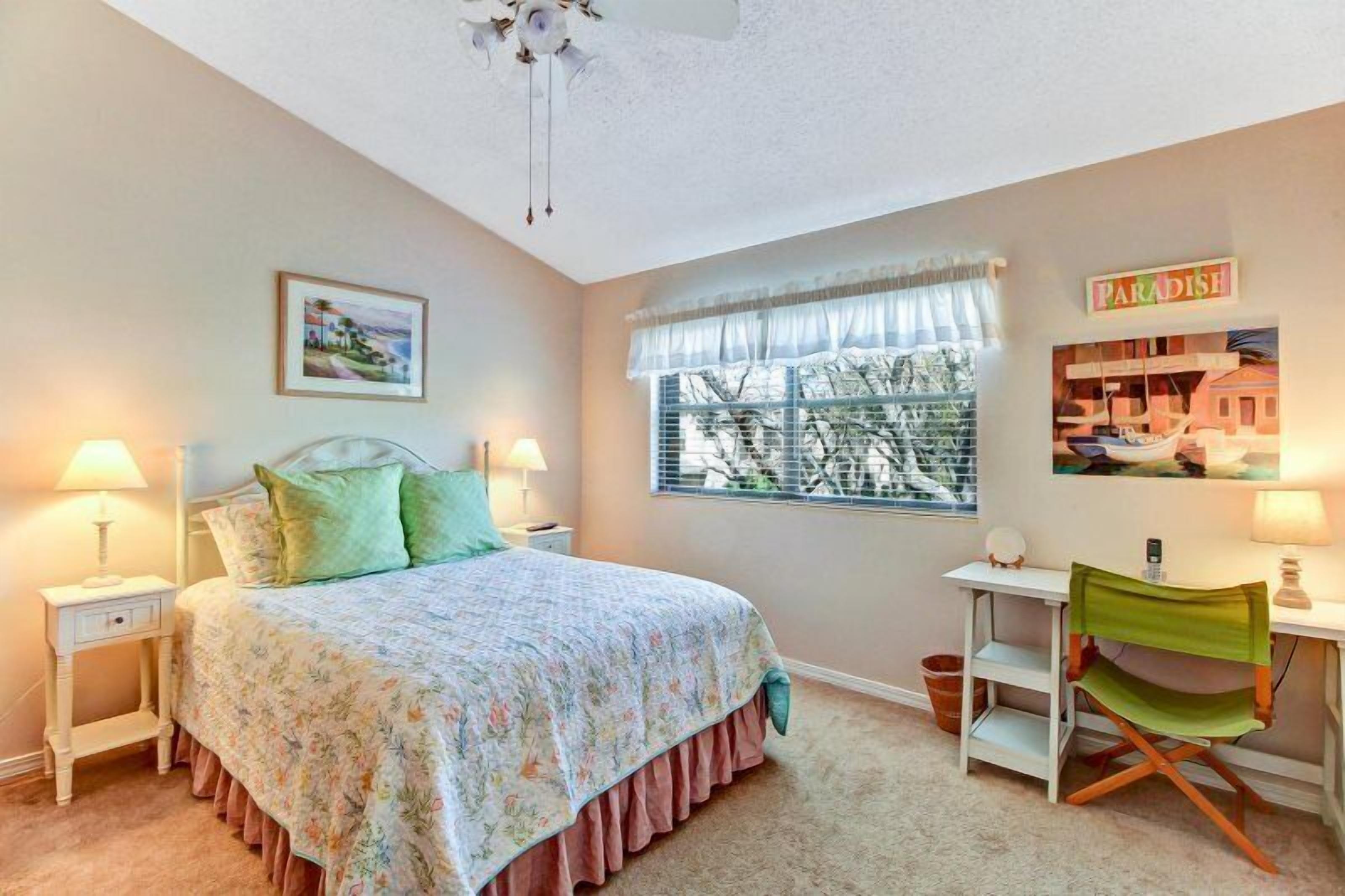 Quarto com cama, duas mesas de cabeceira, escrivaninha, cadeira, janela com persianas, ventoinha no teto e um enfeite de parede com a inscrição 'PARADISE'.