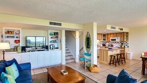 A spacious living room with a sofa, coffee table, and a TV. A kitchen with a dining area and a staircase is visible in the background. - 1109-beach-walker (Fernandina Beach)