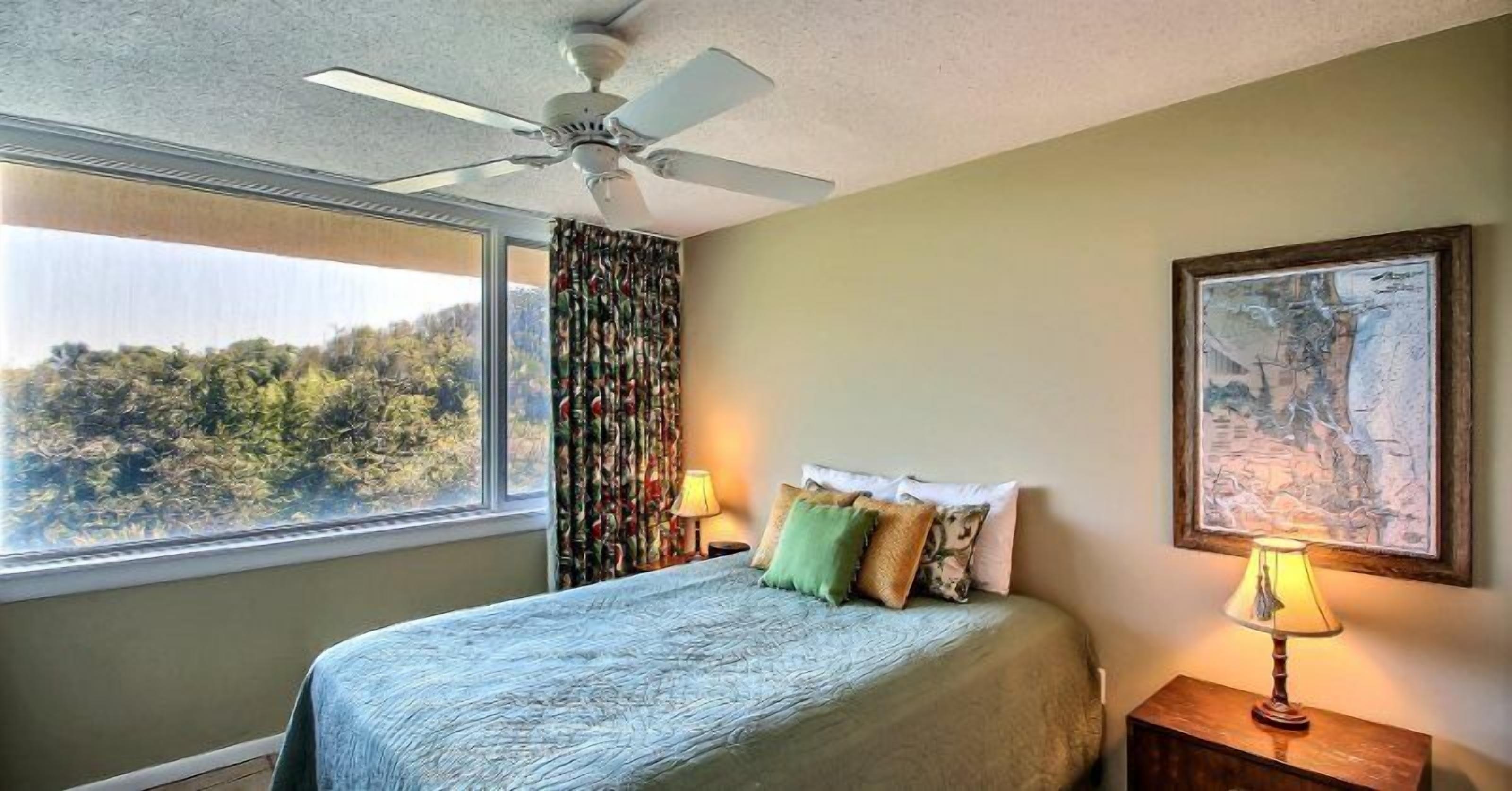 Une chambre à coucher avec un lit, un ventilateur de plafond, une fenêtre avec des rideaux et un tableau au mur.