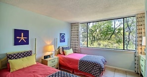 A bedroom with two beds, a nightstand, a window with a view of trees, and wall art. - 1109-beach-walker (Fernandina Beach)