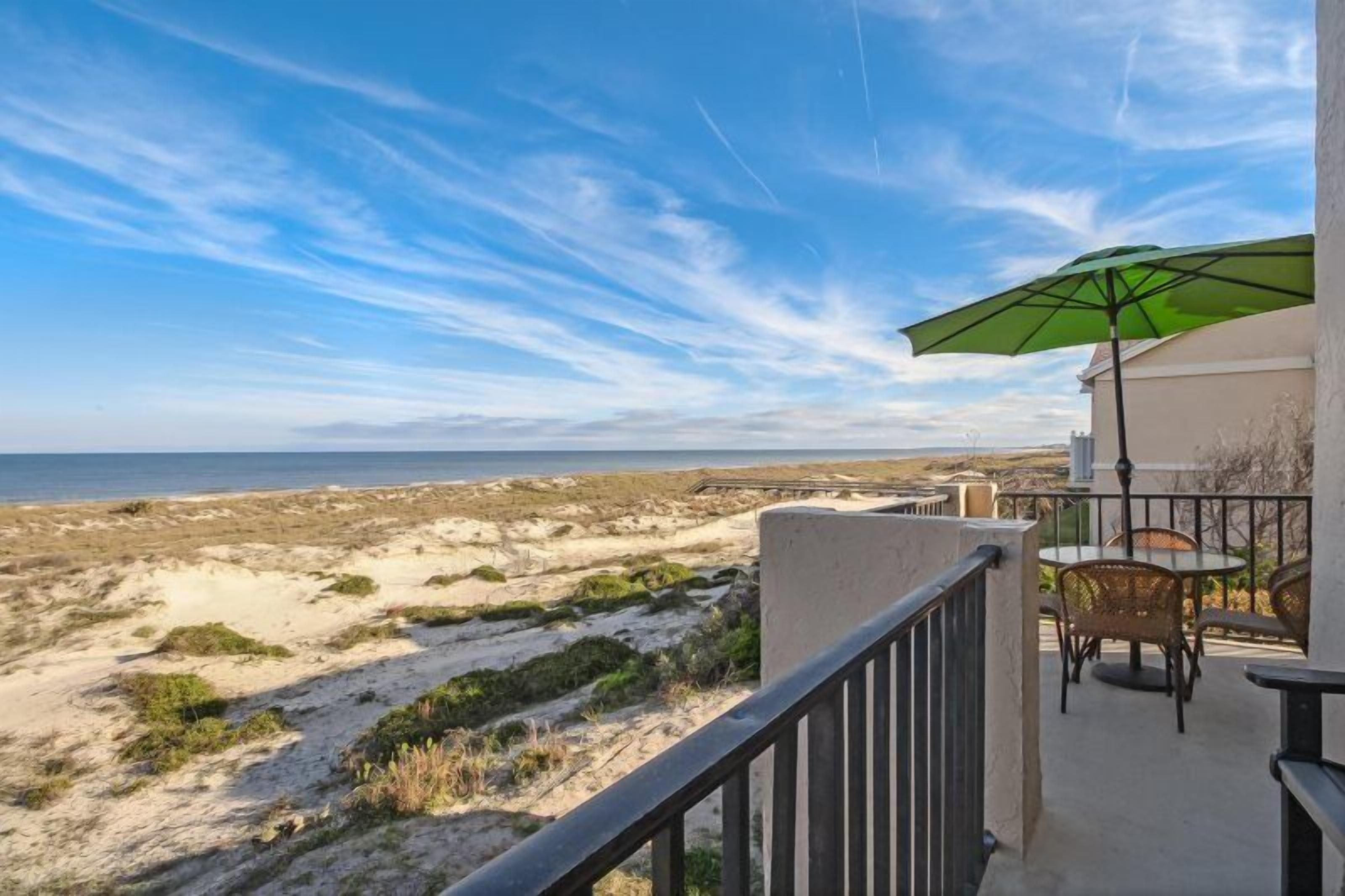 Uma varanda com vista para a praia, uma mesa com cadeiras e um guarda-sol verde.