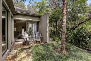A modern house with a glass door entrance, a covered patio with outdoor furniture, and a lush garden with various plants and a palm tree.