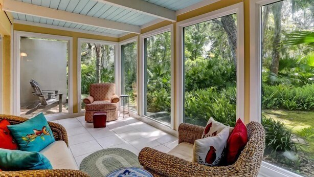 A sunlit room with large windows, wicker furniture, and a view of lush greenery outside.
