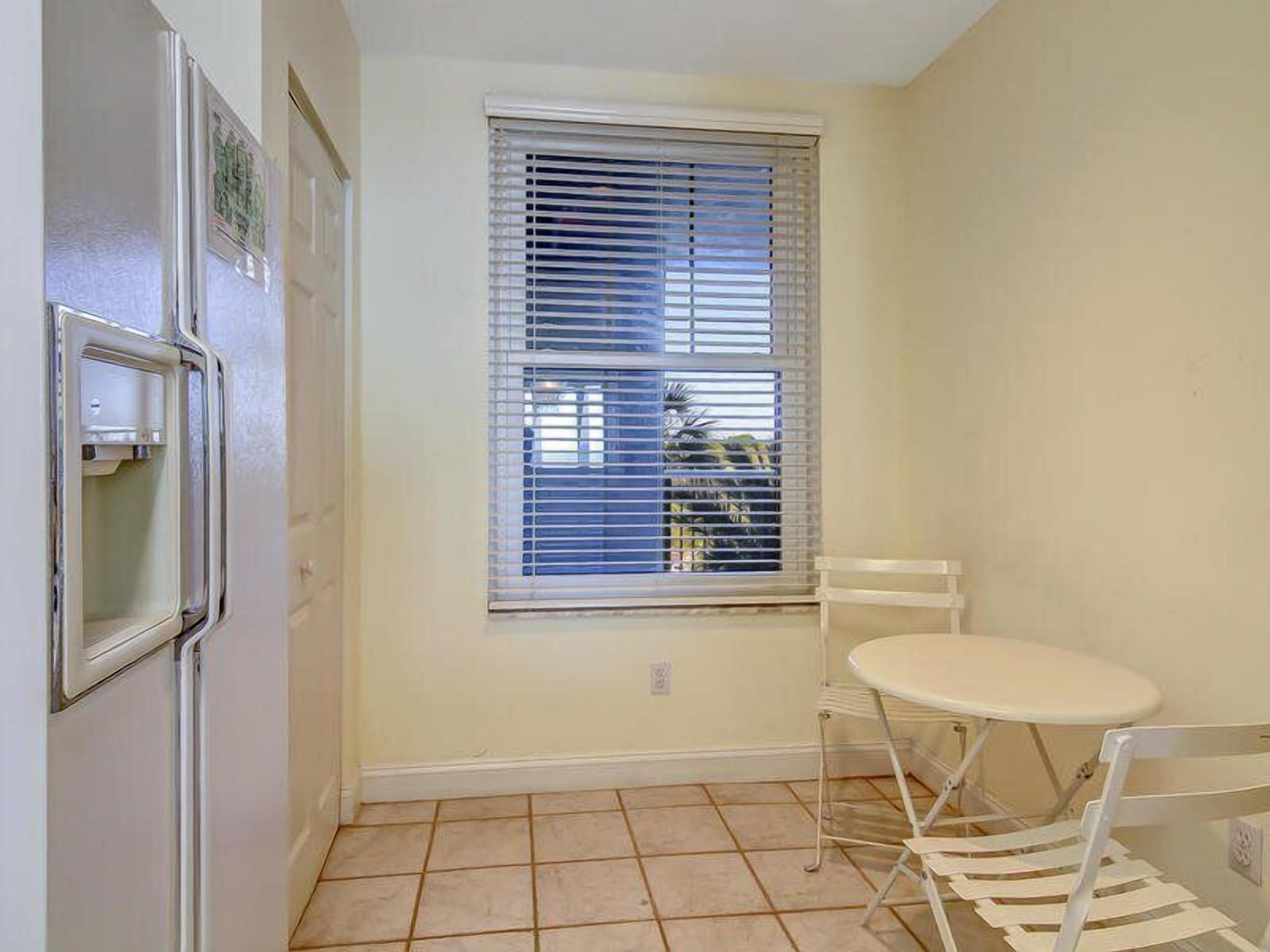 A compact kitchen with a refrigerator, a window with blinds, a small table, and a chair.