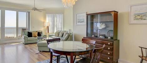 A spacious living room with a round dining table, two sofas, a chandelier, and a large window overlooking the ocean.