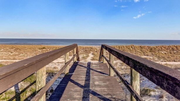 Een houten looppad dat naar een strand leidt, met duinen en een heldere lucht.