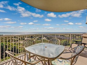 Un balcon avec une table et des chaises en verre, donnant sur un paysage côtier sous un ciel dégagé.