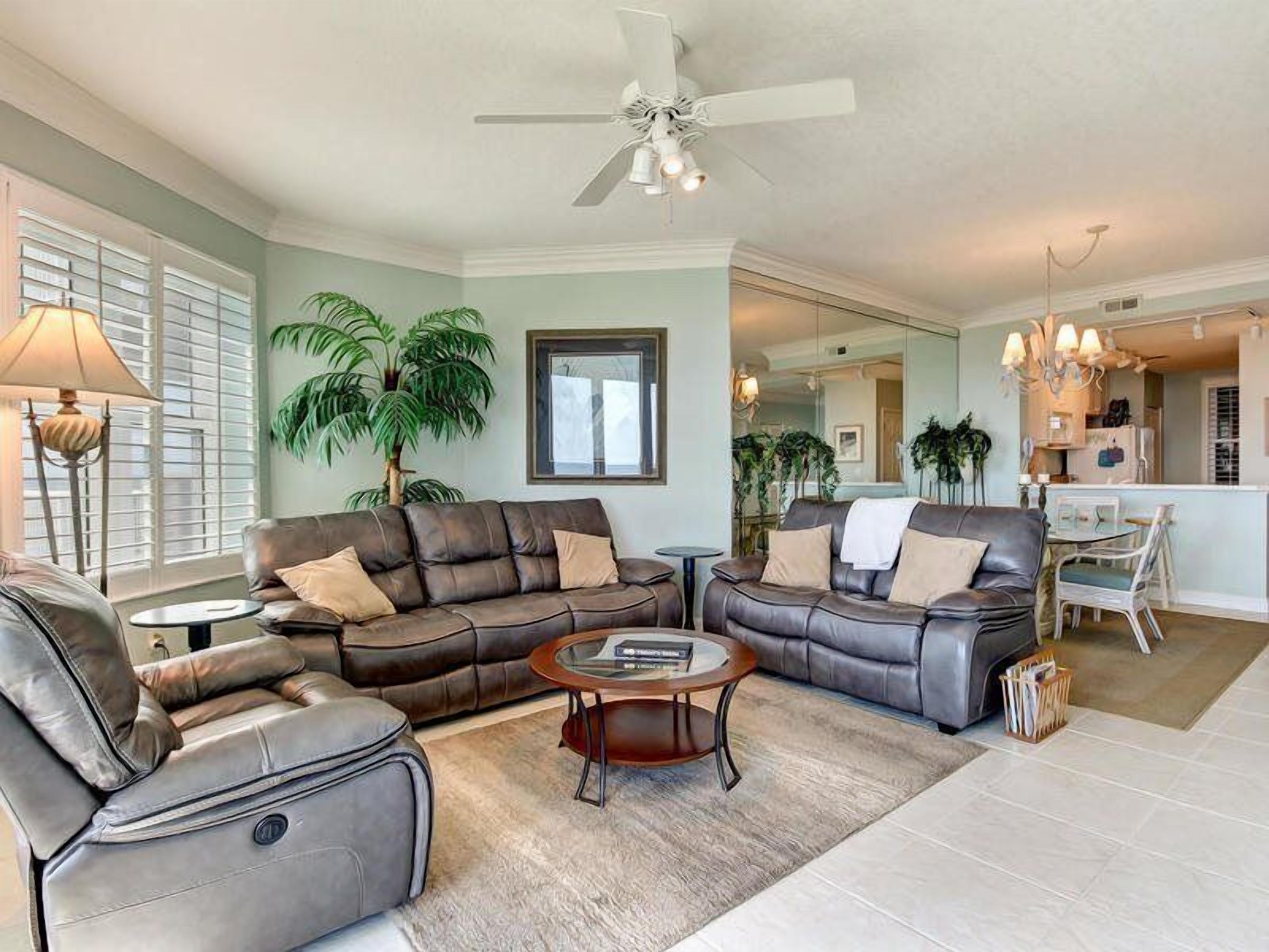 A living room with a sofa, armchair, and coffee table. There is a ceiling fan, a lamp, and a plant.