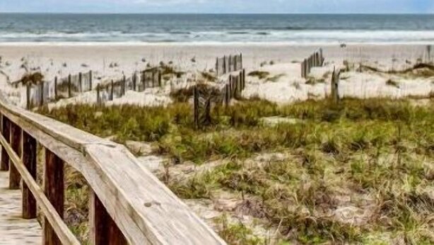 Ein hölzerner Steg führt zu einem Strand mit Dünen und spärlicher Vegetation.