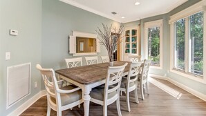 A dining room with a wooden table and white chairs, a built-in cabinet with decorative plates, and large windows with a view of greenery. - 13-green-winged-teal (Fernandina Beach)