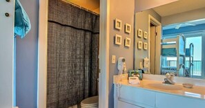 Une salle de bain avec un grand miroir, un lavabo et un rideau de douche. Des tableaux encadrés ornent le mur et une serviette est accrochée à la porte.