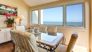 Un comedor con mesa de vidrio, sillas acolchadas, vista al mar y un cuadro en la pared.