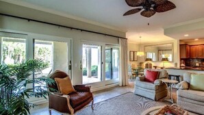 A spacious living area with a large window, a ceiling fan, and a kitchen area visible through the glass doors. - 1801-turtle-dunes (Fernandina Beach)