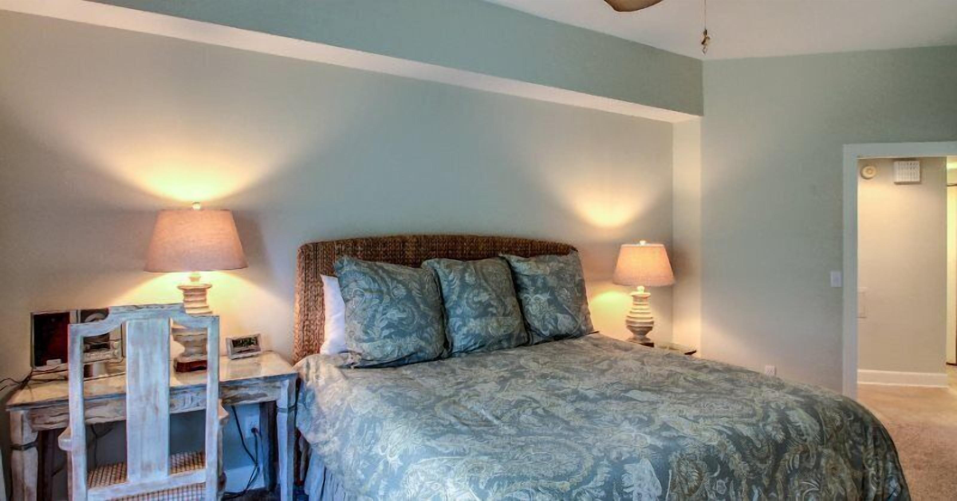 Quarto com cama, duas mesas de cabeceira com abajures, uma cadeira e uma escrivaninha.