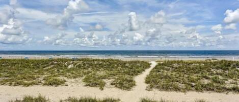 Une plage avec un sentier de sable menant à l’océan, sous un ciel partiellement nuageux.