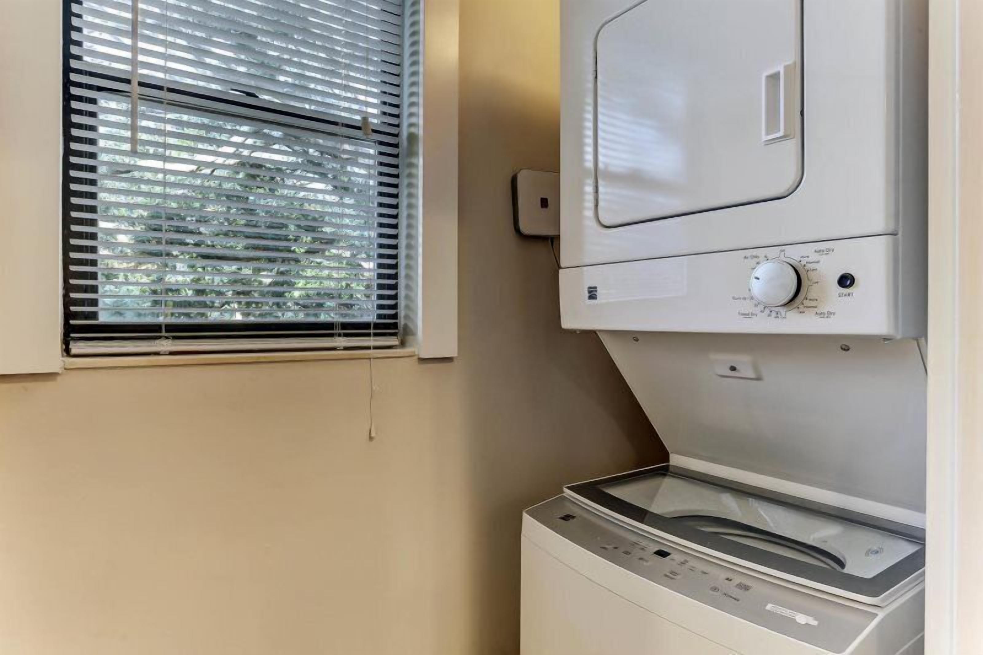 A laundry room with a washing machine and dryer, a window with blinds, and a wall-mounted electrical outlet.