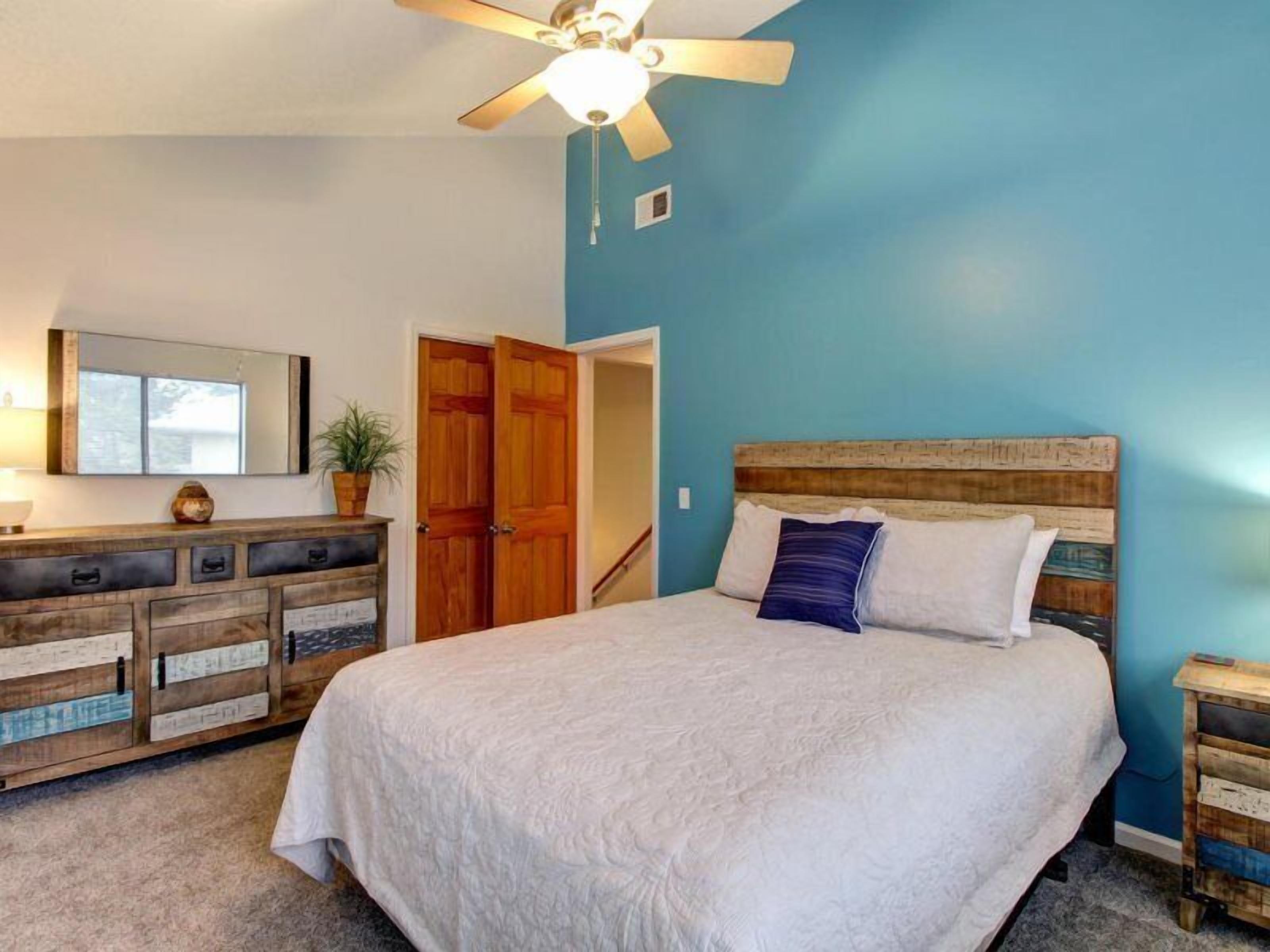 Un dormitorio con una cama grande, cabecera de madera, ventilador de techo, cómoda con varios cajones y una puerta que da a otra habitación.