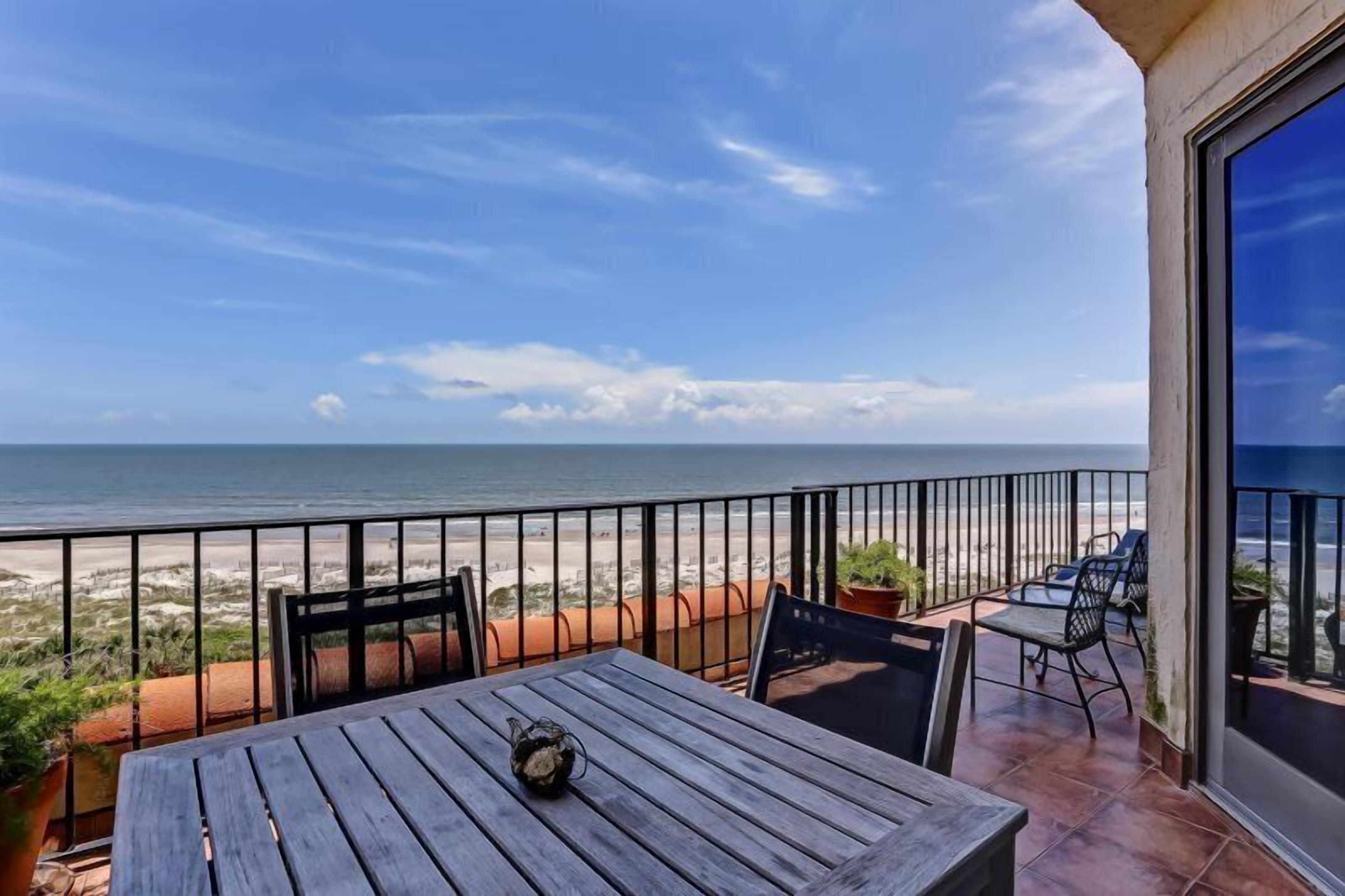 A balcony with a view of the beach, a table, chairs, and potted plants.