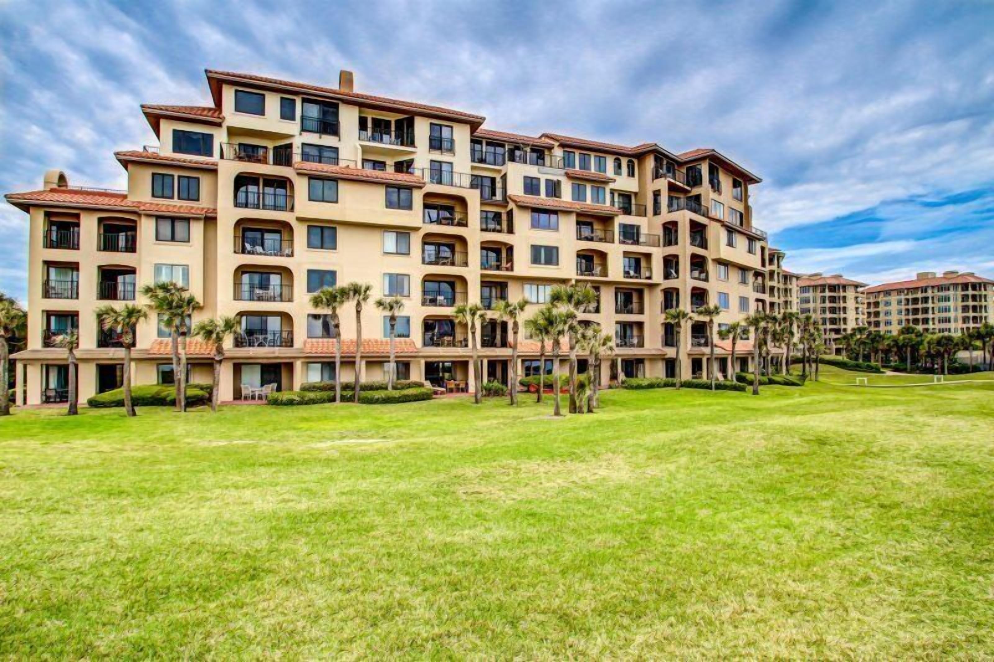 A multi-story hotel with balconies, a well-maintained lawn, and palm trees.