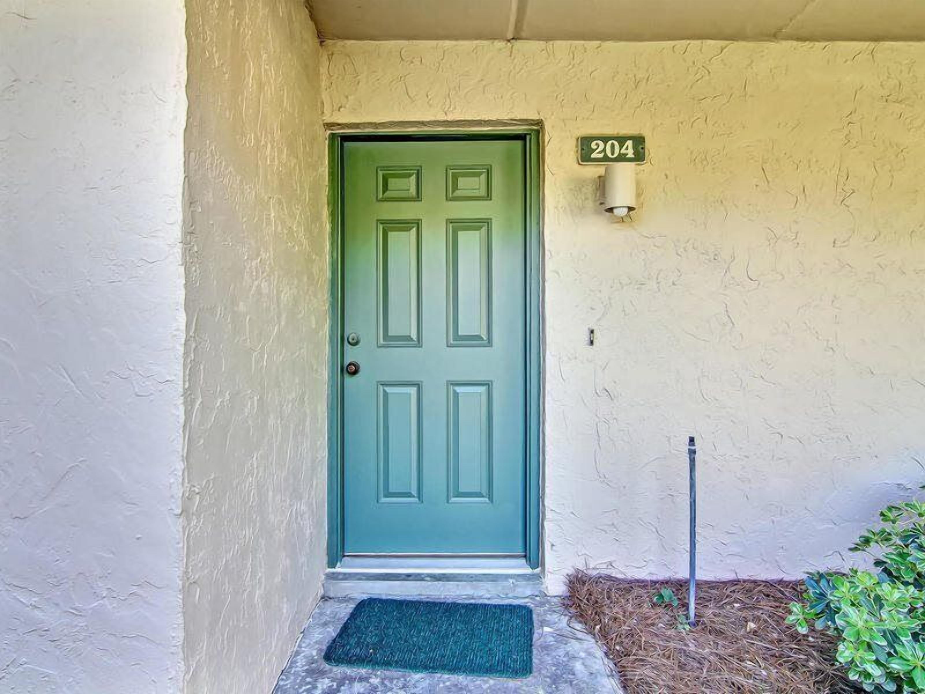 Une porte verte avec une plaque portant le numéro « 204 » au-dessus, un petit paillasson à l’entrée et un luminaire fixé au mur.