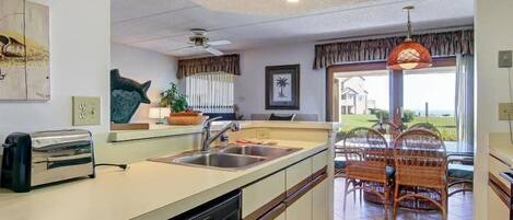 A kitchen with a dishwasher, a toaster, and a dining area with wicker chairs and a view of the outdoors.