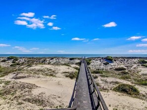 Un malecón de madera que lleva a una playa con dunas y vegetación escasa.