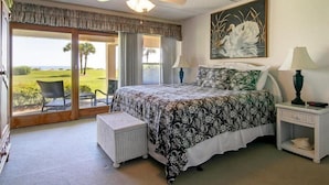 A bedroom with a large bed, a ceiling fan, a painting, a nightstand, and a view of a golf course through a sliding door. - 204-sailmaker (Fernandina Beach)