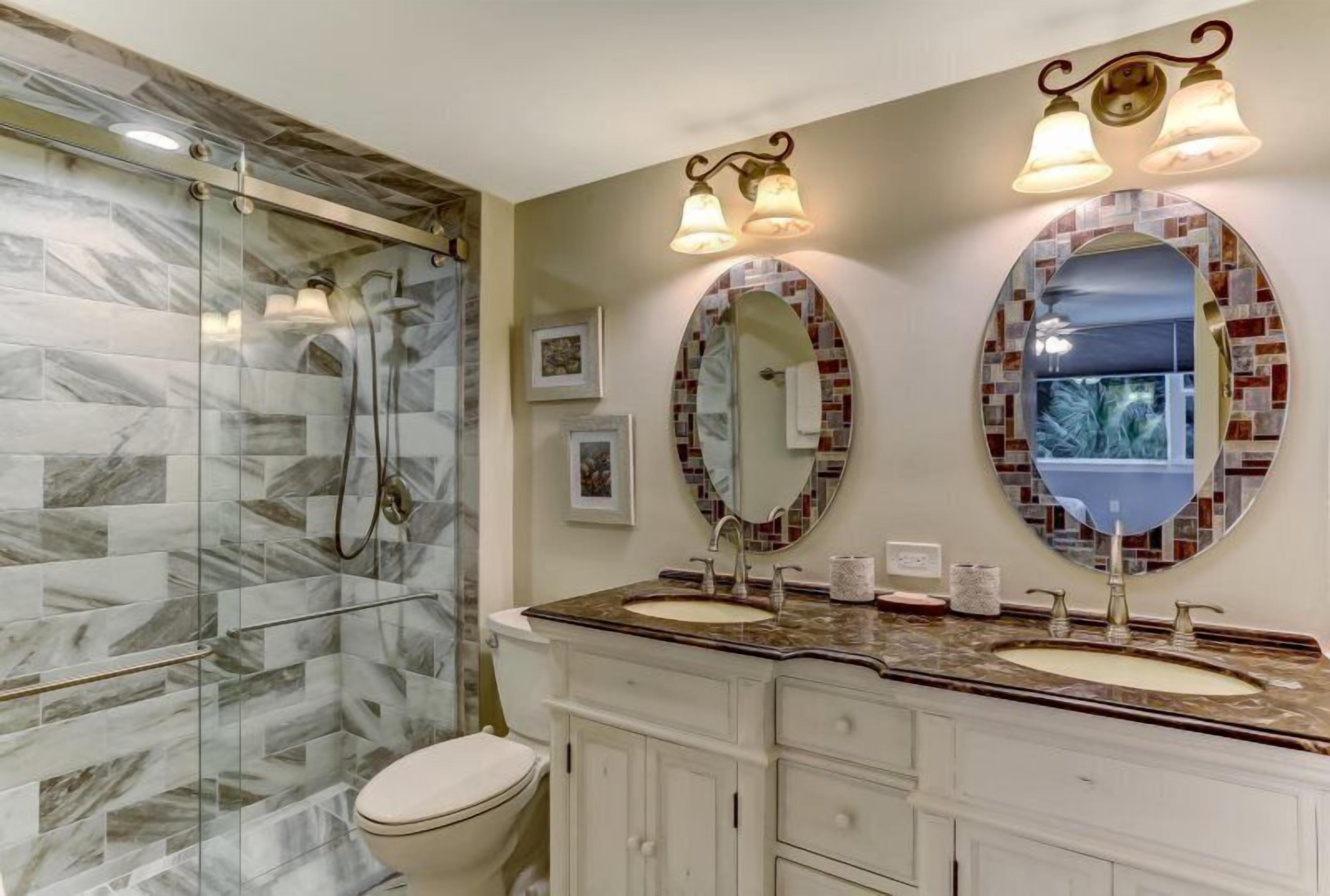 A bathroom with a glass shower enclosure, two sinks with vanity cabinets, and two wall-mounted mirrors with light fixtures.