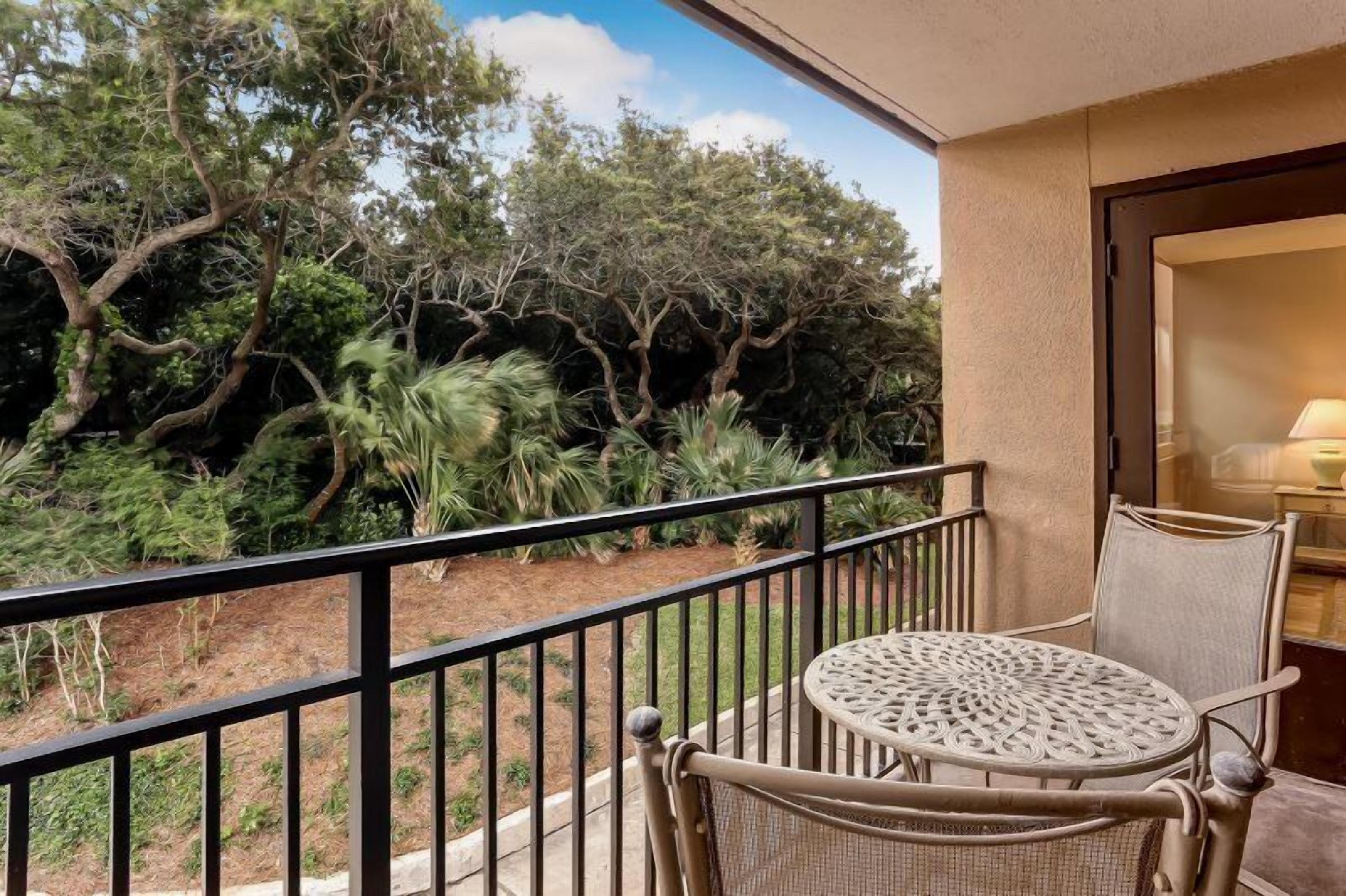 A balcony with wicker furniture, a view of trees, and a door leading to a room with a lamp.